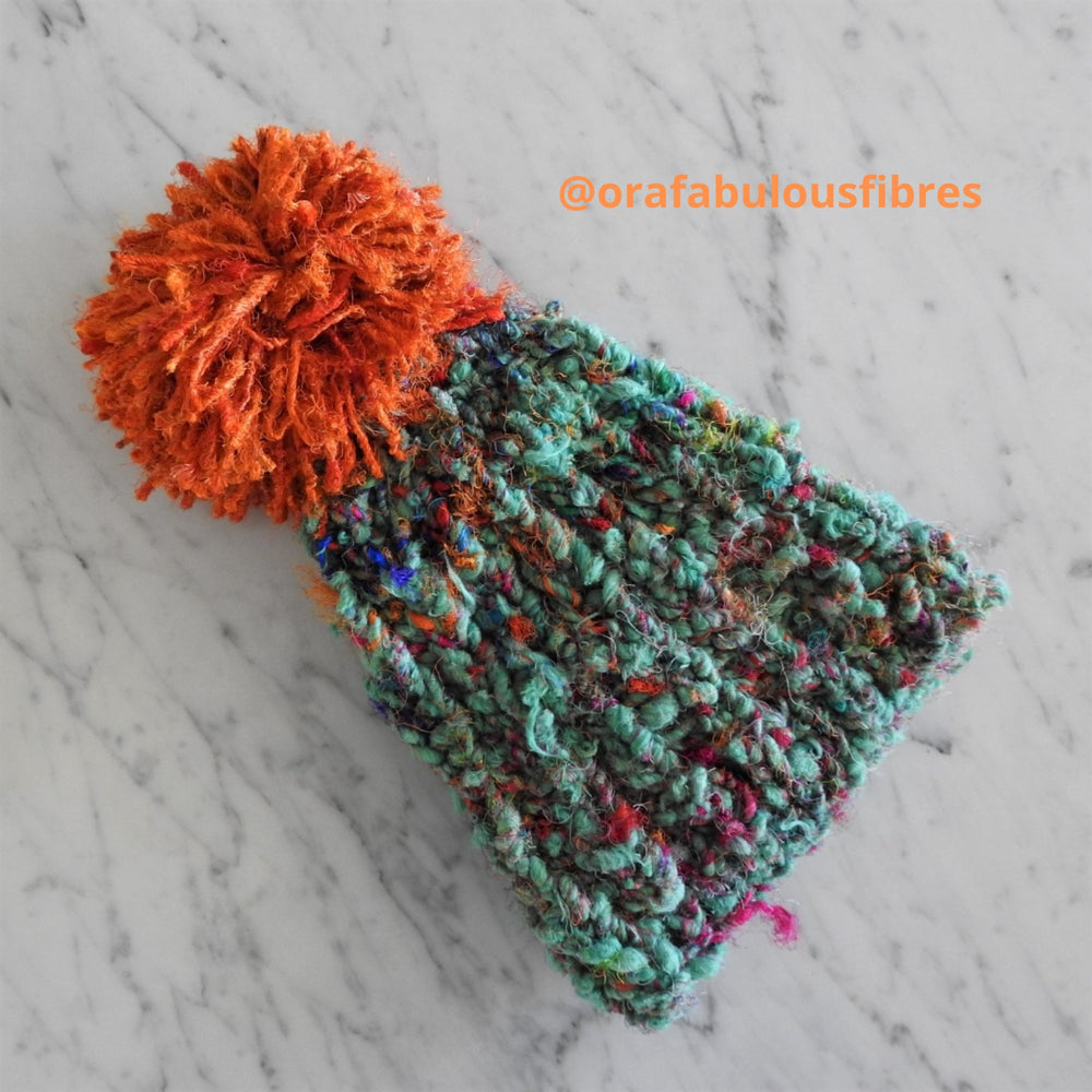 Knitted beanie using chunky wool and silk recycled yarn in green. Beanie has a large, soft pom pom made using orange Recycled Sari Silk yarn. Hat is knitted in brioche stitch